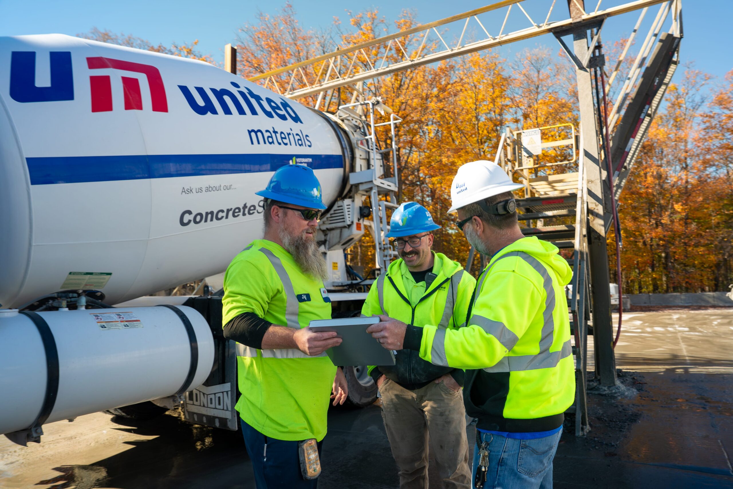 three male workers in hard hats and PPE standing and talking in front of a United Materials truck, while reviewing work materials, in Orchard Park, Buffalo, Western New York.